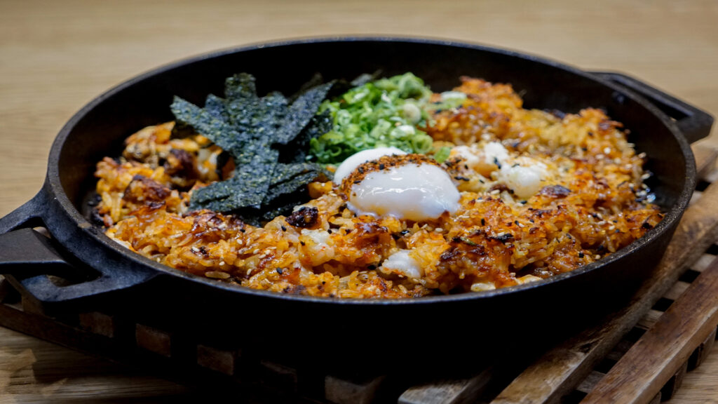 A sizzling dish of crispy fried rice topped with a soft poached egg, green onions, and seaweed in a cast-iron skillet.