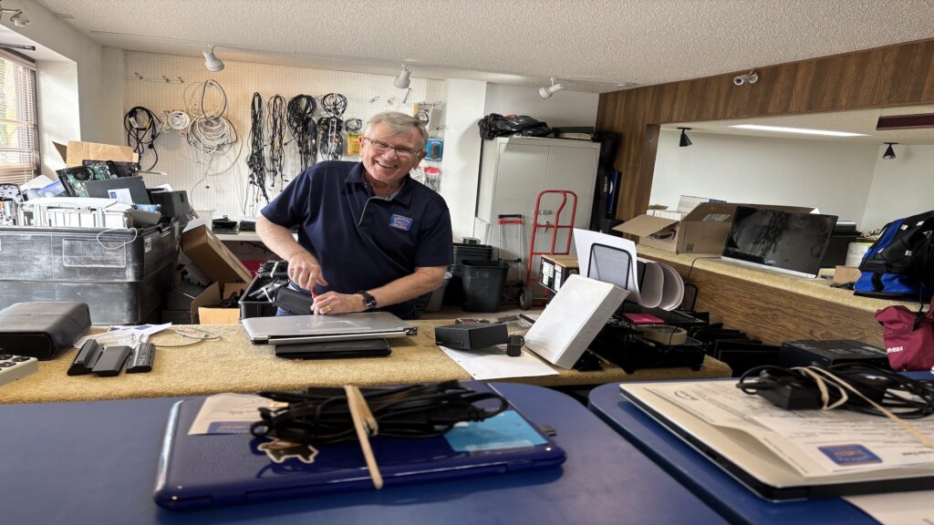 A cheerful man working on a laptop at a counter in a workshop filled with cables and electronic equipment.