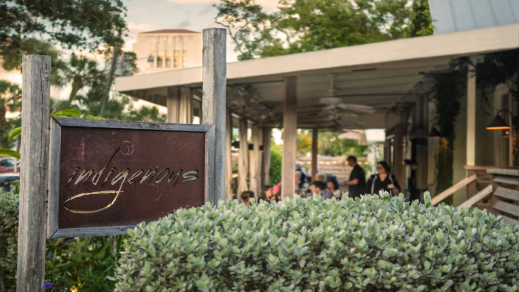 bush partially covering a brown restaurant sign that spells "indigenous".