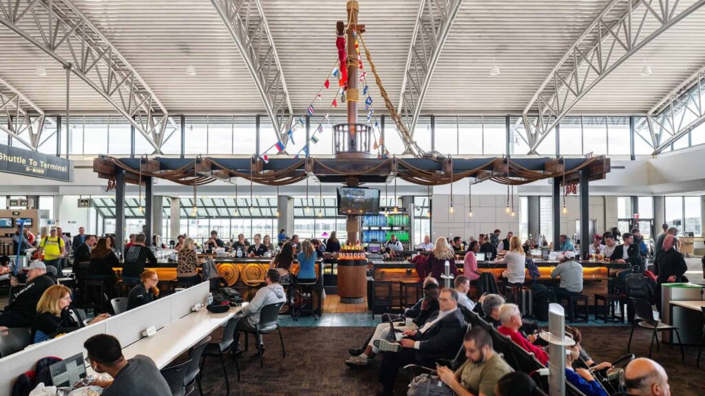 a large number of travelers gather outside gates at an airport, other enjoy a drink at a pirate ship themed bar